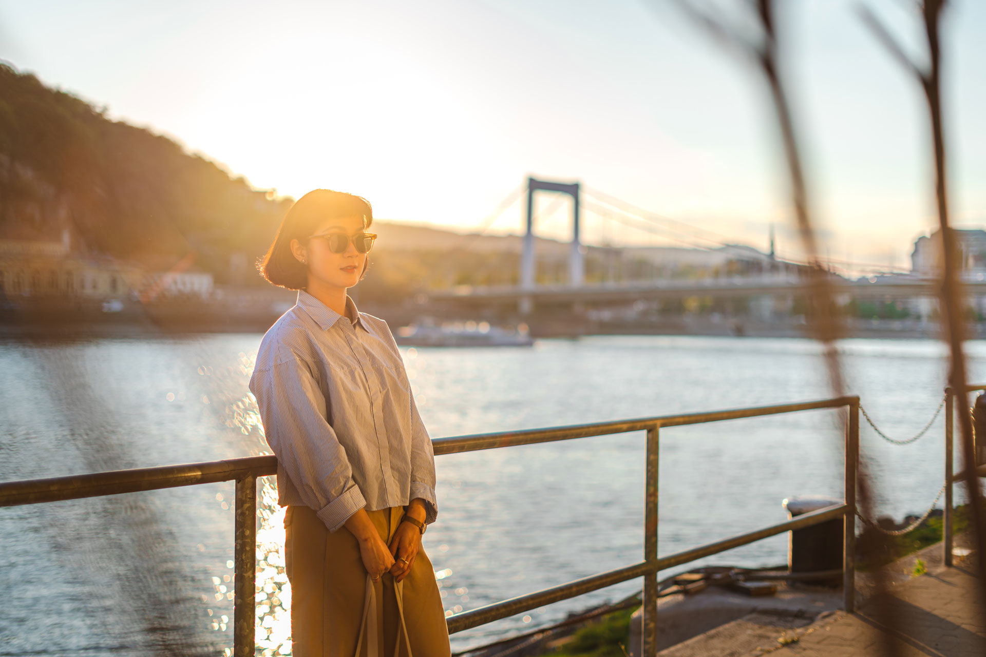 A woman with short dark hair and sunglasses, wearing a blue shirt and khaki pants, stands by a metal railing at sunset. Behind her is the Danube River and the illuminated white Elizabeth Bridge in Budapest