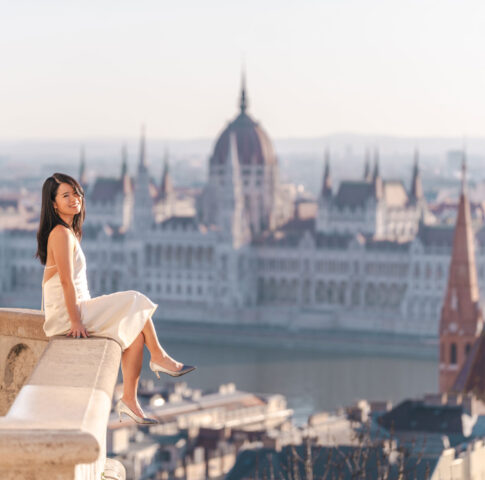 A smiling woman in a white silk dress sitting on a stone balcony at Fisherman's Bastion, with the Hungarian Parliament Building and the Danube River blurred in the background in Budapest.