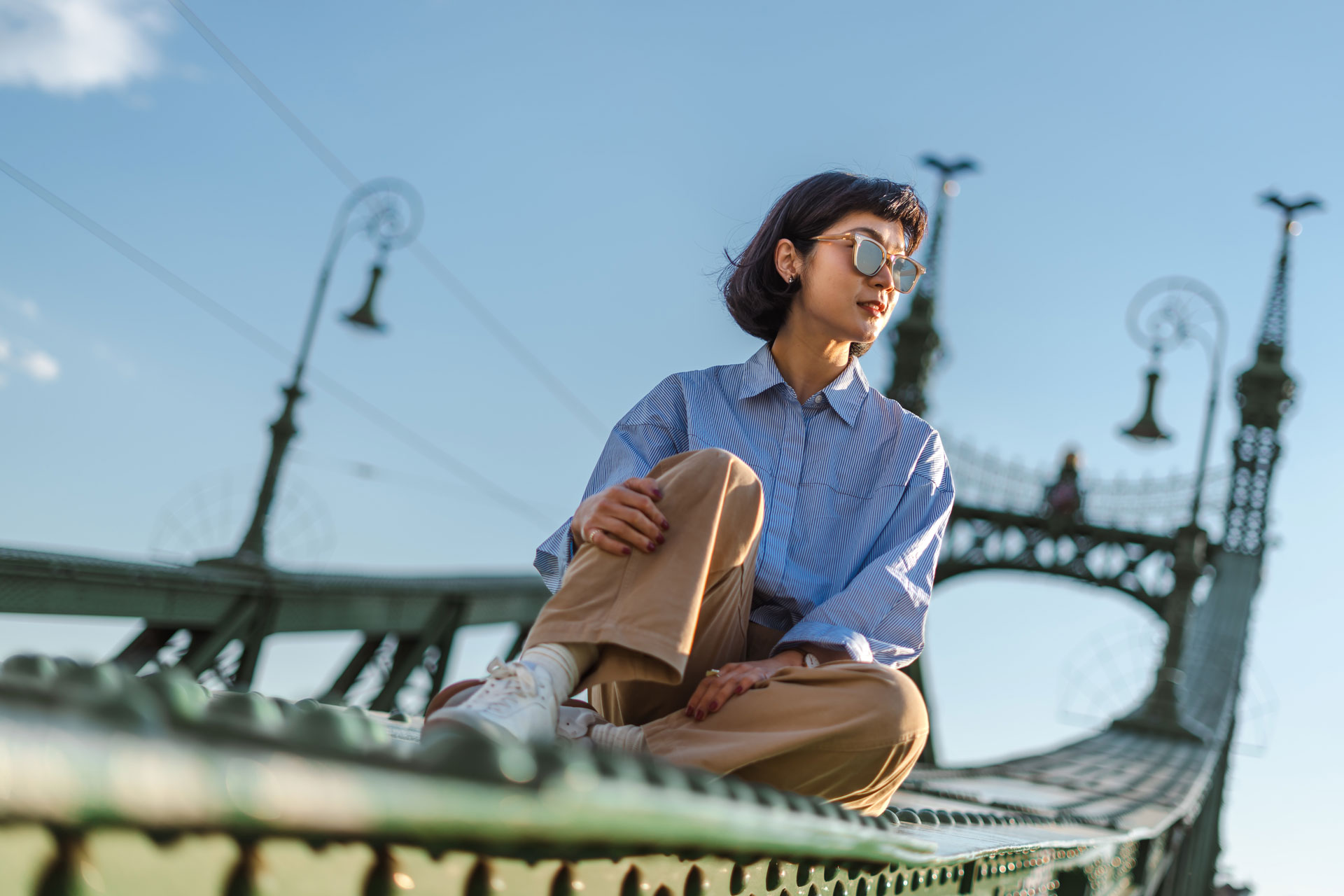 A woman with short dark hair wearing sunglasses and a blue striped shirt, sitting on the green iron structure of the Liberty Bridge in Budapest during golden hour