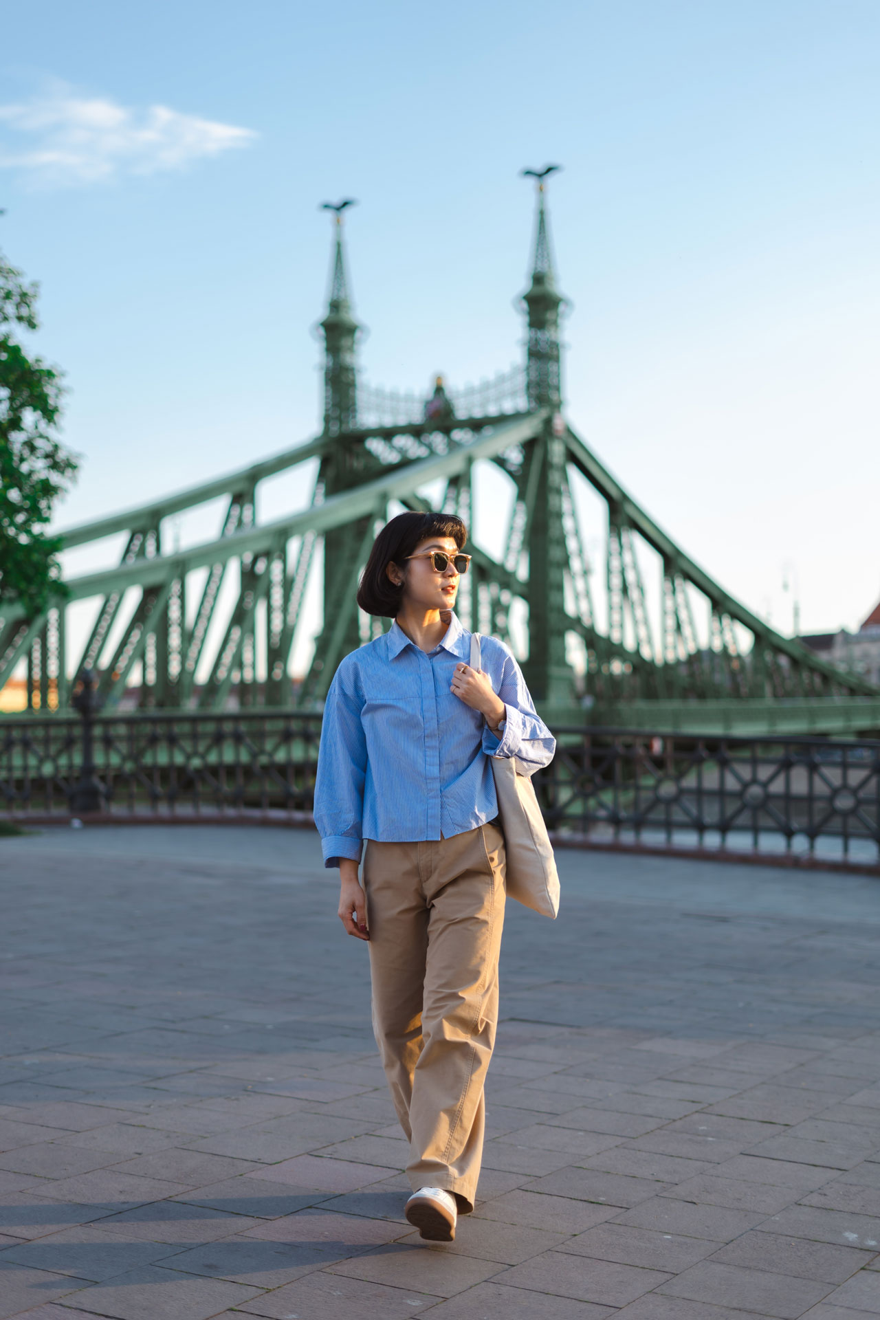 A woman in a blue striped shirt and khaki pants walking with a tote bag in front of the green Liberty Bridge in Budapest during the afternoon sun