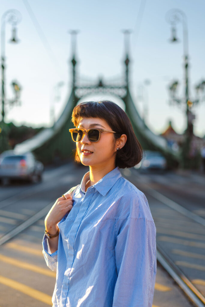 A portrait of a woman with a dark bob haircut, wearing sunglasses and a blue striped shirt. She is holding a tote bag strap and looking to the side. Behind her, the ornate green metal structure of the Liberty Bridge in Budapest is softly in focus with golden light